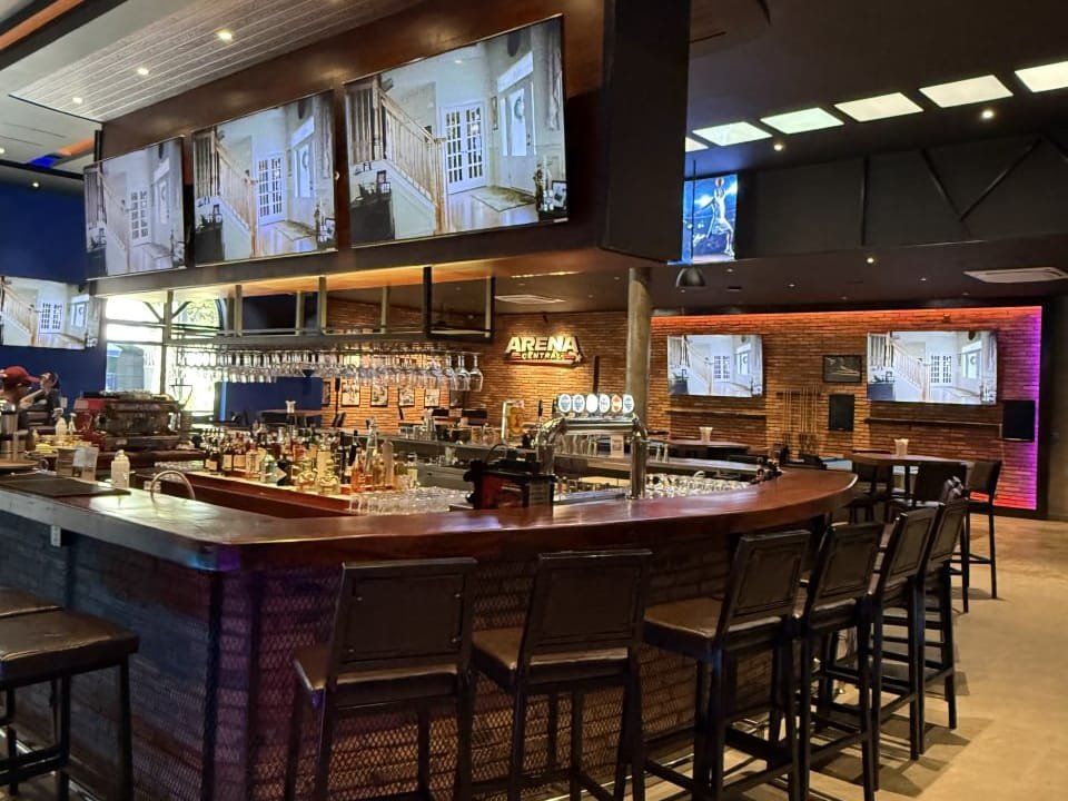 Curved island bar under the lit Arena Central sign, with live sport on the screens above